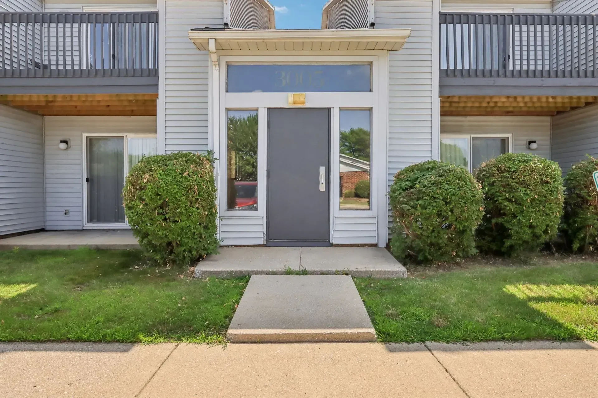 A tan, two-story apartment building exterior with a central grey entry door, green shrubs, and upper-level balconies.