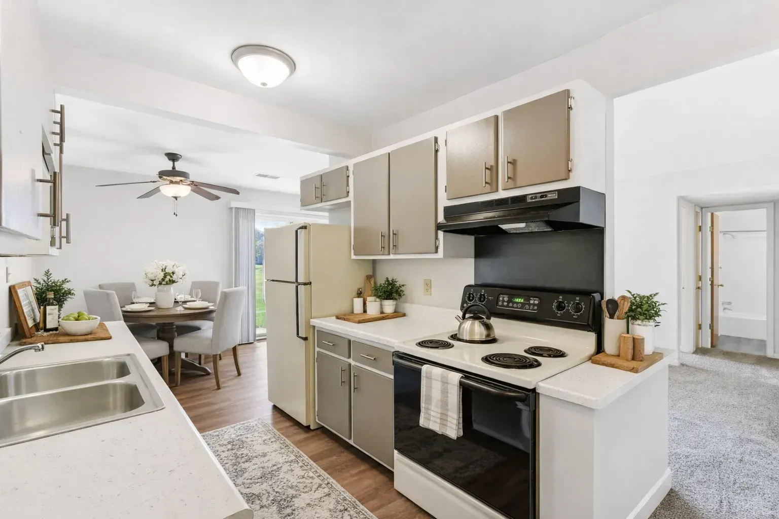 A bright kitchen with light-colored countertops, grey cabinets, a stainless steel stove, and a dining area in the background.
