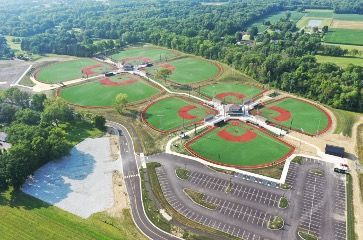 Aerial view of a complex with five baseball fields arranged around central hubs, adjacent to parking and green forest.
