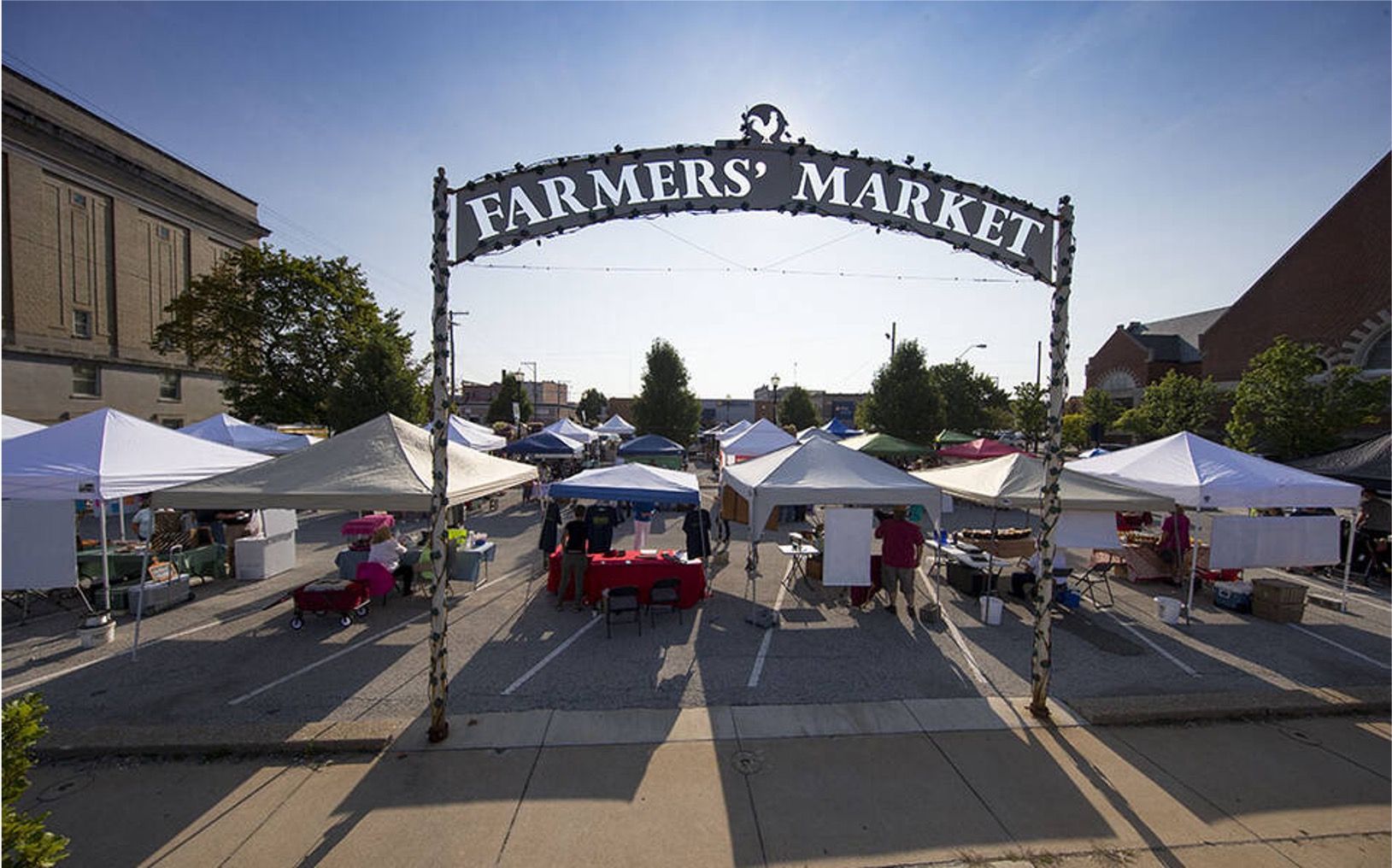 A metal archway labeled "Farmers' Market" opens into a sunny outdoor market filled with white vendor tents.