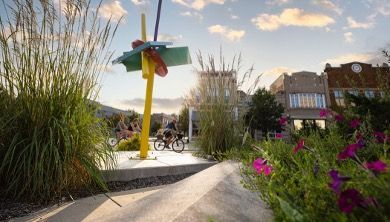 A bright yellow abstract sculpture stands in a sunlit public square, with cyclists nearby and historic buildings beyond.