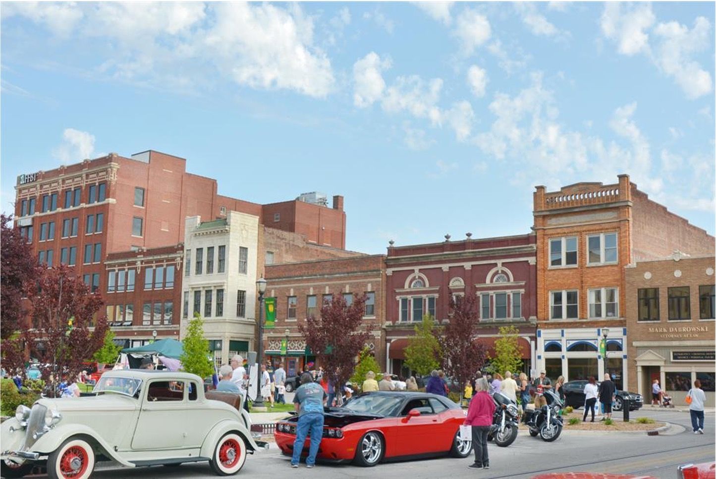 People gather for a car show featuring vintage and modern vehicles parked along a historic brick downtown street.