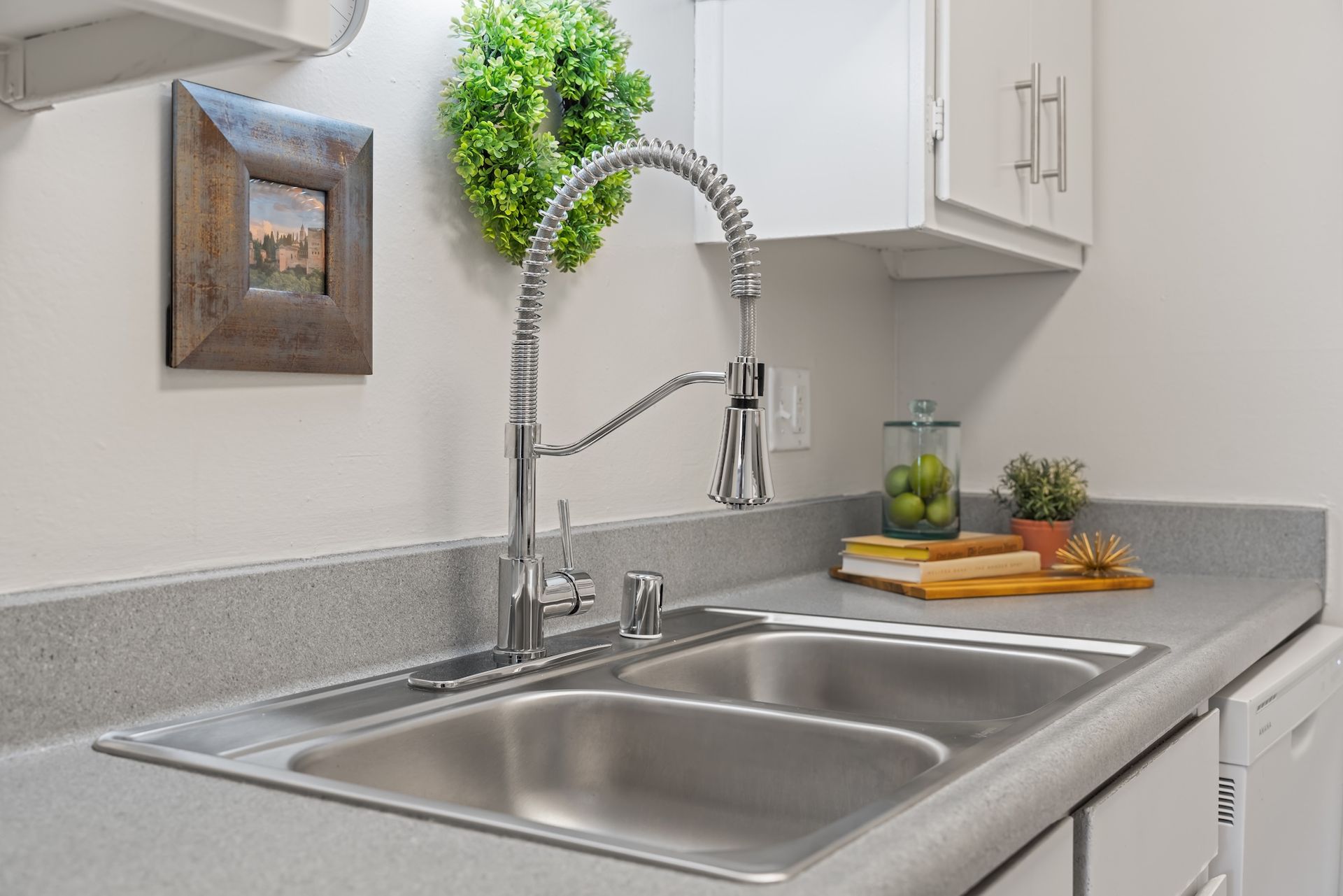 Kitchen sink with chrome faucet, countertop, and decor, including a green wreath and framed art.