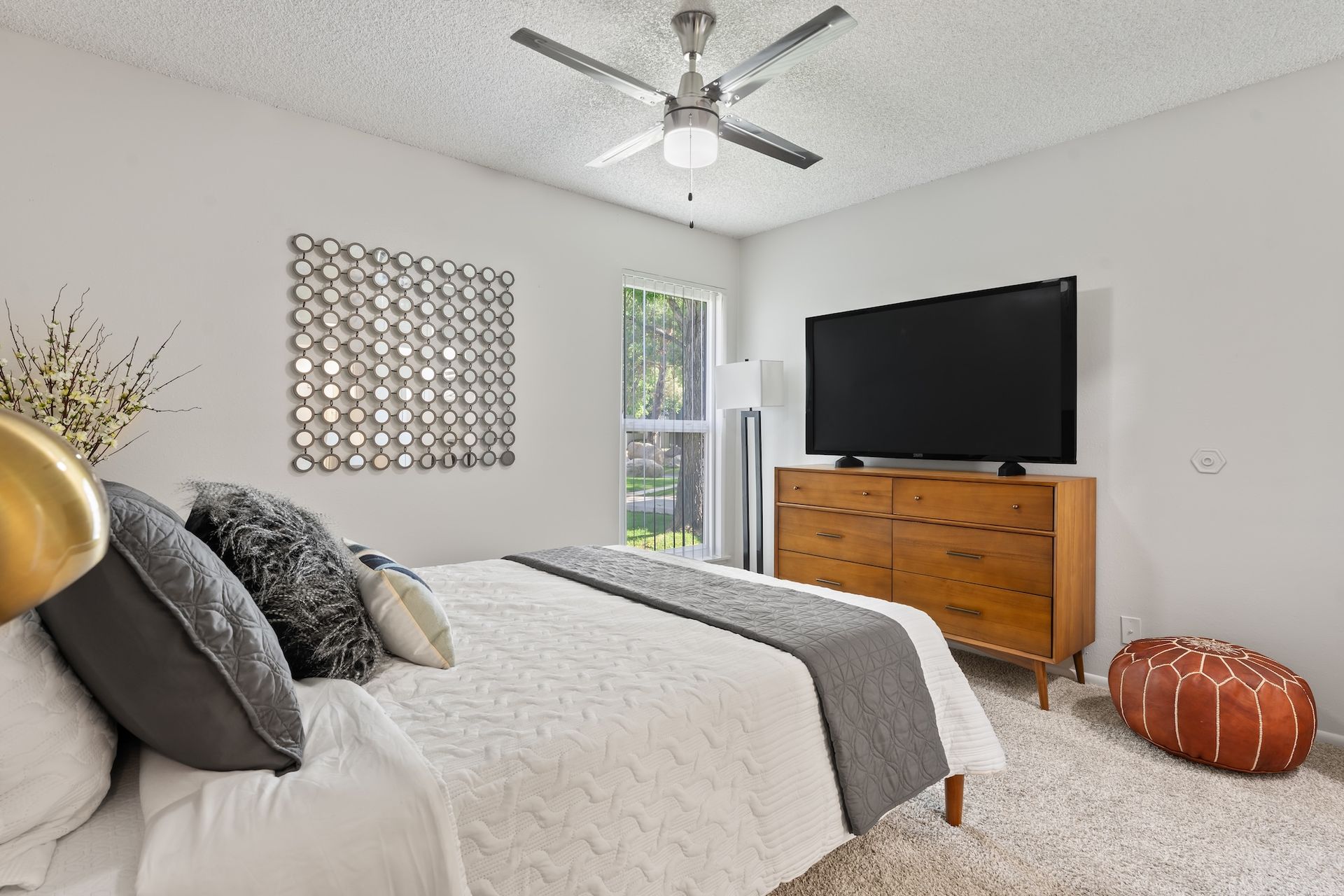Bedroom with bed, dresser, TV, mirror art, and a brown leather pouf on a shag carpet.