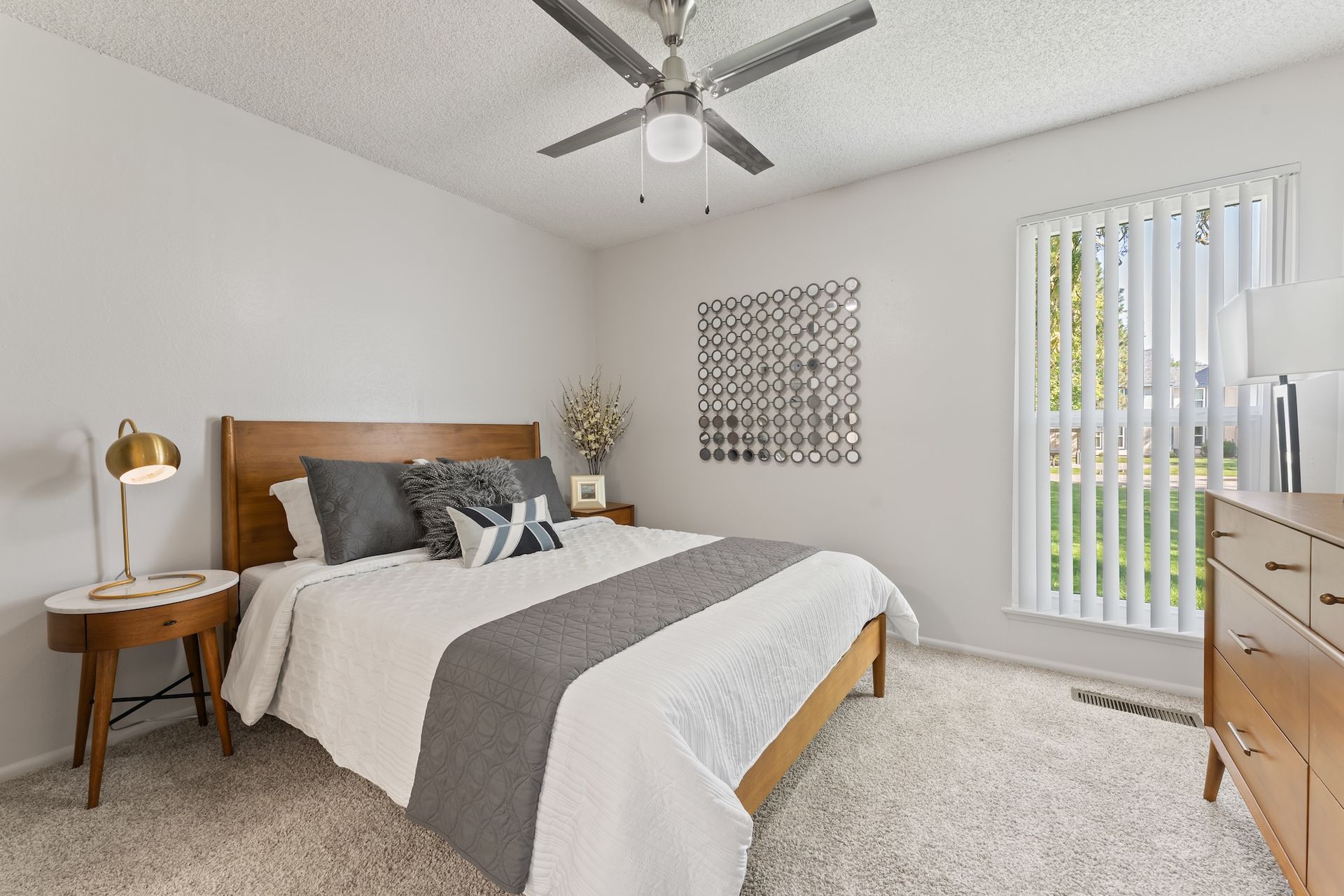 Bedroom with a wooden bed, nightstand, dresser, and white carpet. Window with vertical blinds.