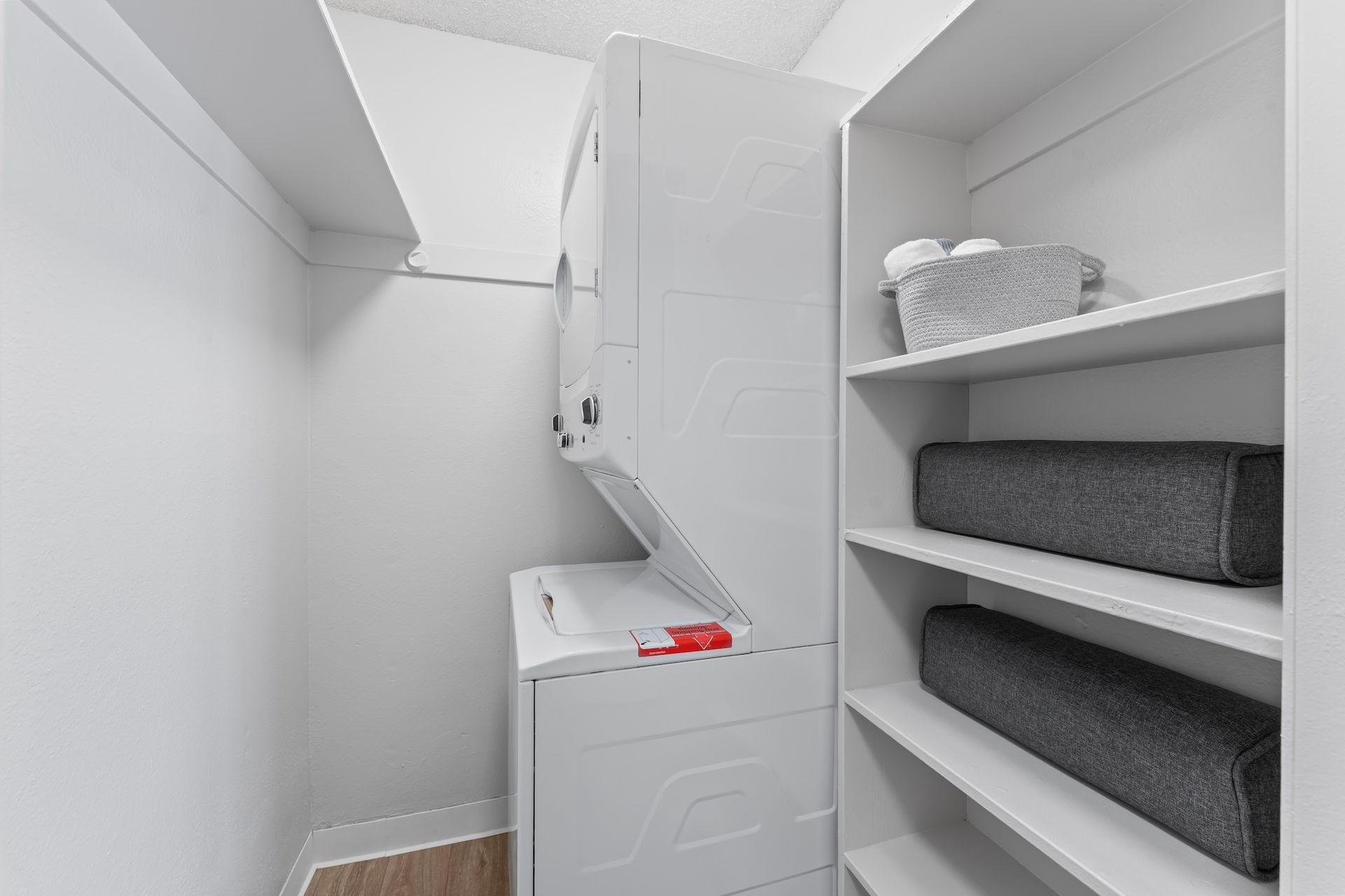 Laundry room with stacked white washer/dryer, shelving holding towels.
