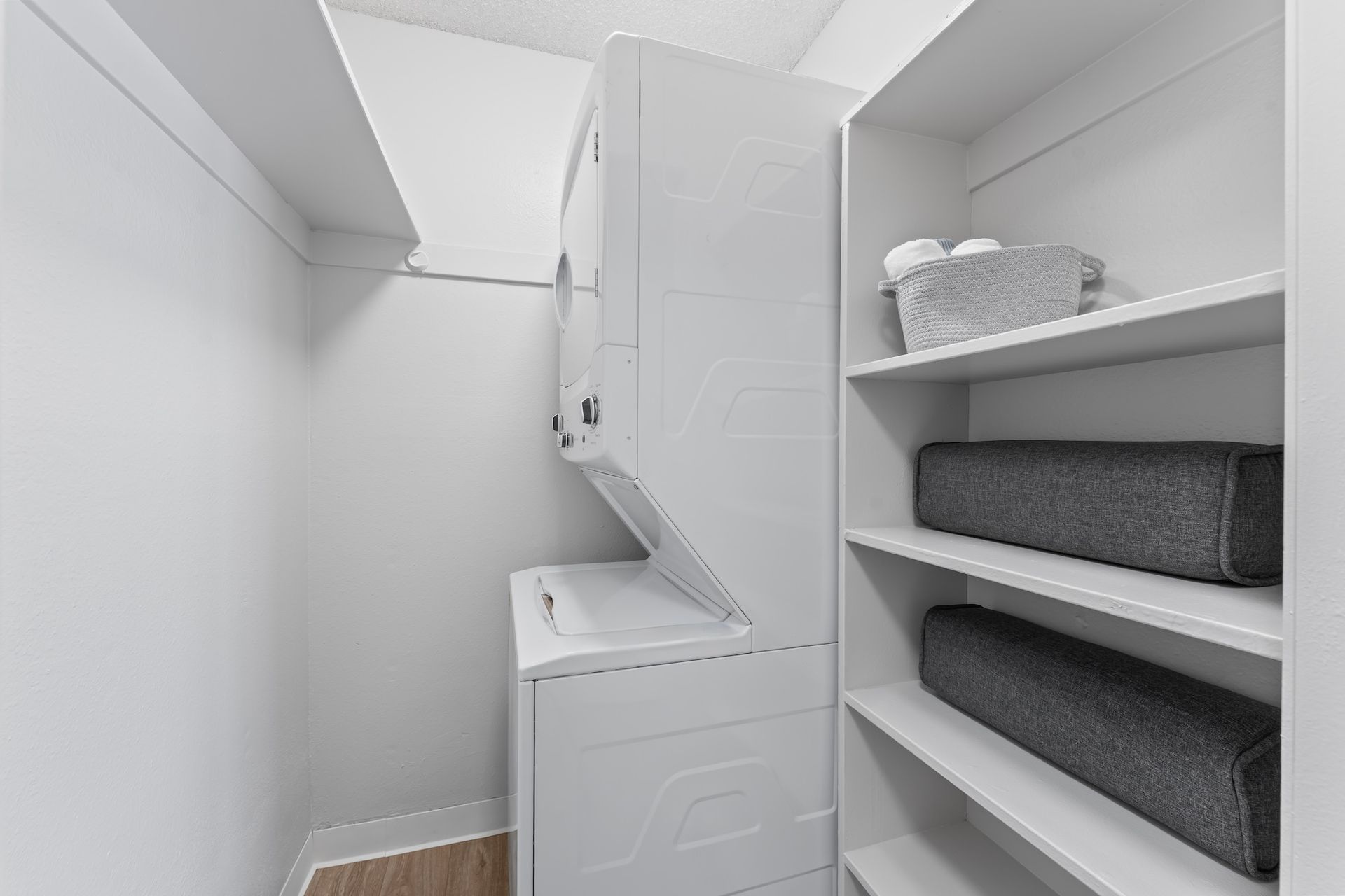 White laundry room with stacked washer/dryer, shelving, and folded gray towels.