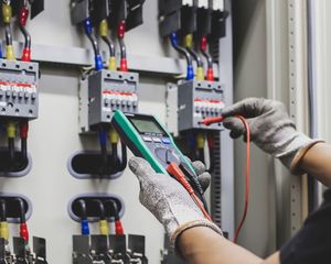 Electrician using a multimeter to test electrical wiring in a panel. Wires are red, blue, and yellow.