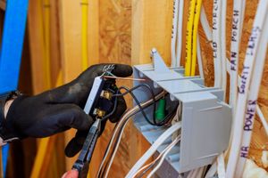 Electrician wiring a switch in a gray electrical box; wood background, black-gloved hands, copper wire, white and yellow cables.