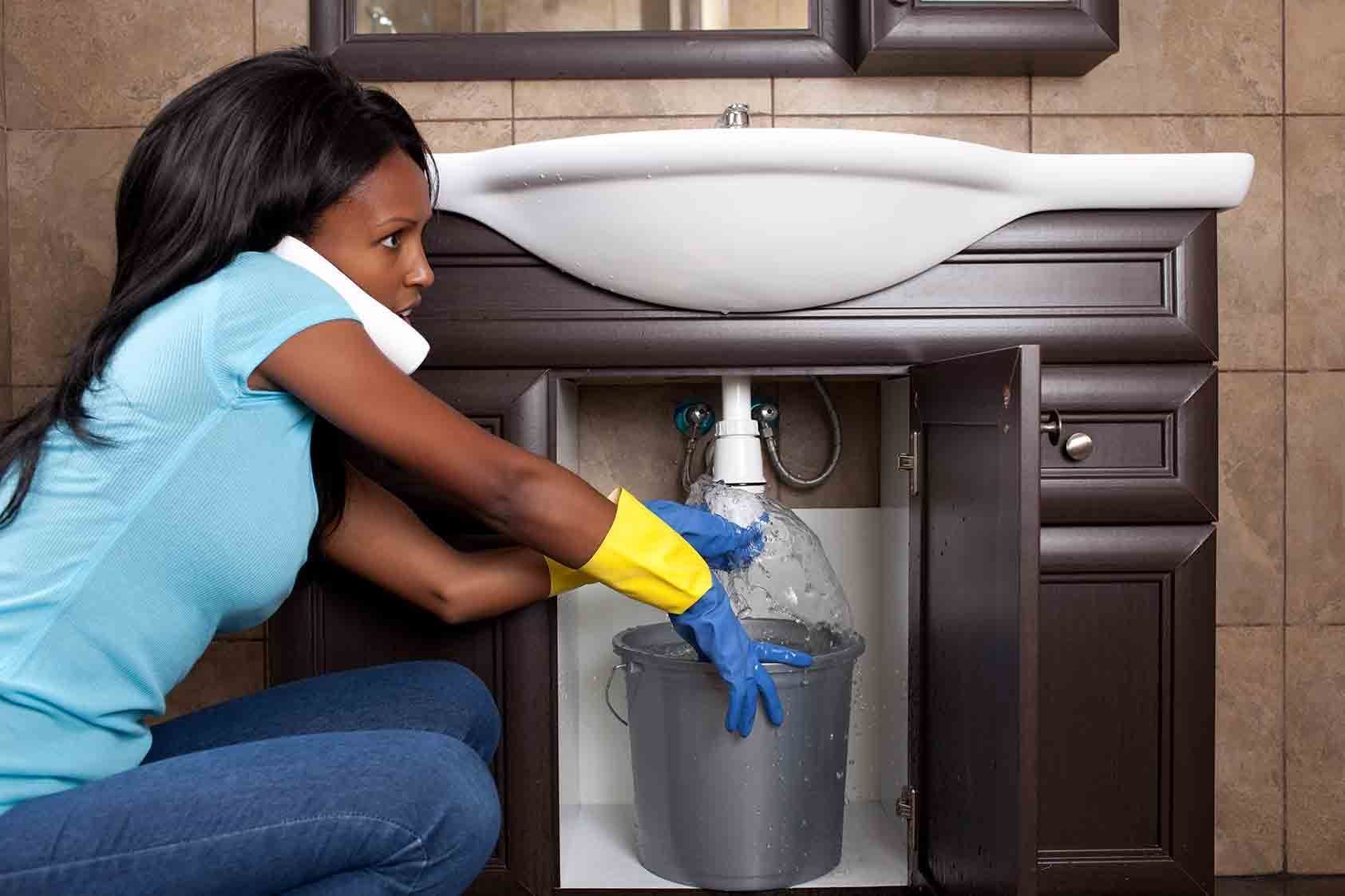 A person wearing gloves holds a bucket under a leaking bathroom sink while talking on a phone tucked between their shoulder.