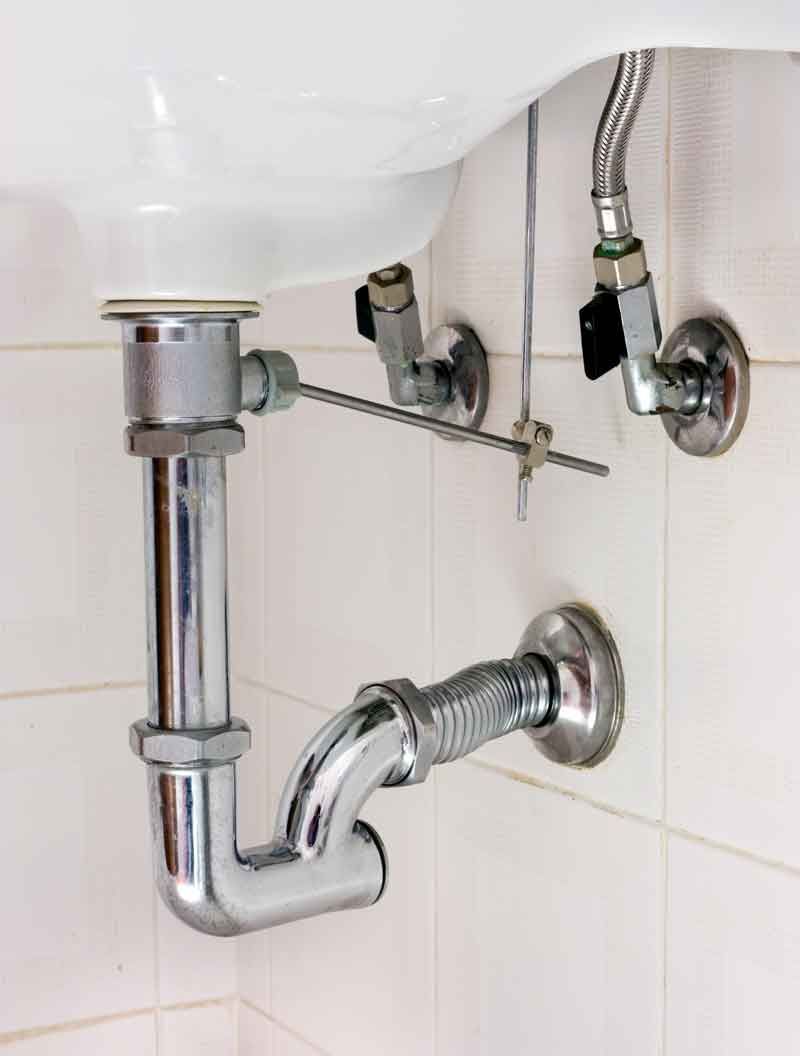 Undersink plumbing with a silver chrome P-trap and two water supply valves mounted on a white tiled wall.