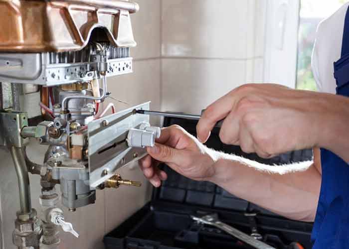 A technician in blue work overalls uses a screwdriver to repair an open gas water heater.