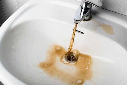 A faucet pours murky, rust-colored water into a white bathroom sink, leaving brown stains around the drain.