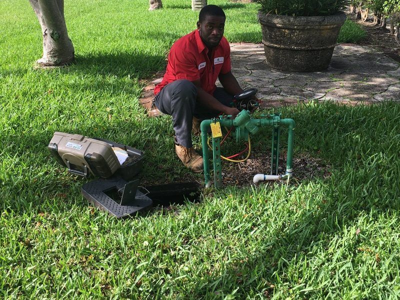 A technician in a red shirt kneels on a lawn, repairing a green irrigation pipe valve system next to a utility case.