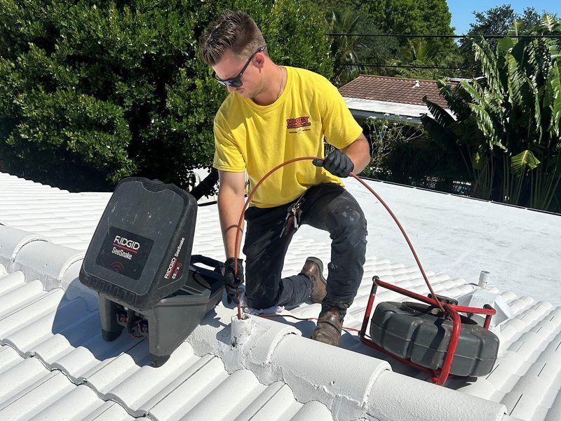 A person in a yellow shirt operates professional pipe inspection equipment on a white tiled roof.