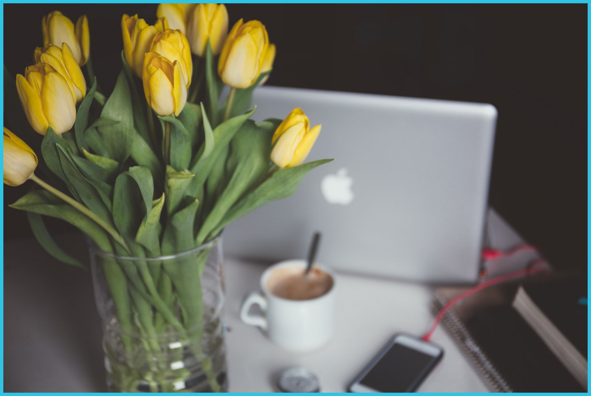 A vase of yellow tulips sits on a table next to a laptop and a cup of coffee.