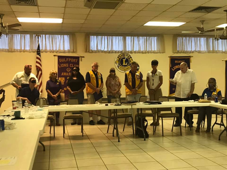 Group of people standing, some wearing vests, in a meeting room with Lions Club banners and an American flag.