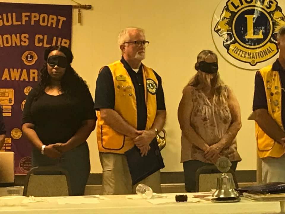 People with blindfolds stand behind a table with a bell in a Lions Club meeting.