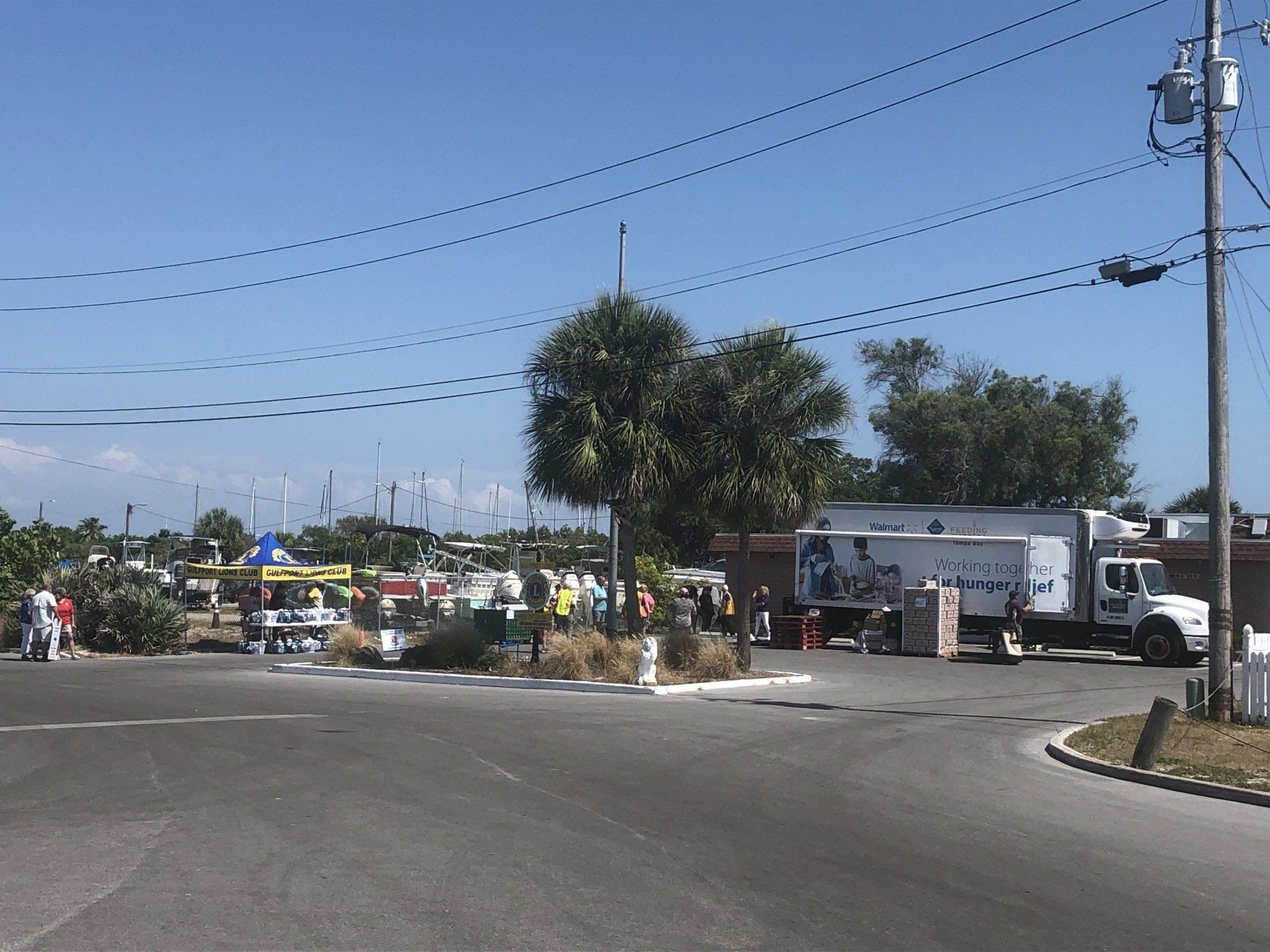 Busy outdoor scene with a commercial truck, people, boats, and businesses under a blue sky.