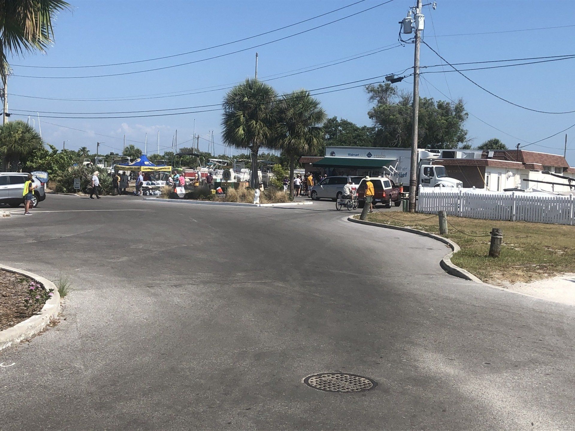 An intersection with people near a building and boats in the background under a blue sky.