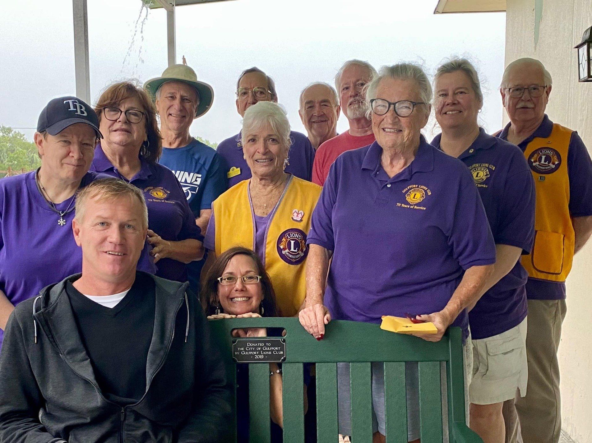 Group of Lions Club members, mostly seniors, smile outdoors. Purple shirts and yellow vests.
