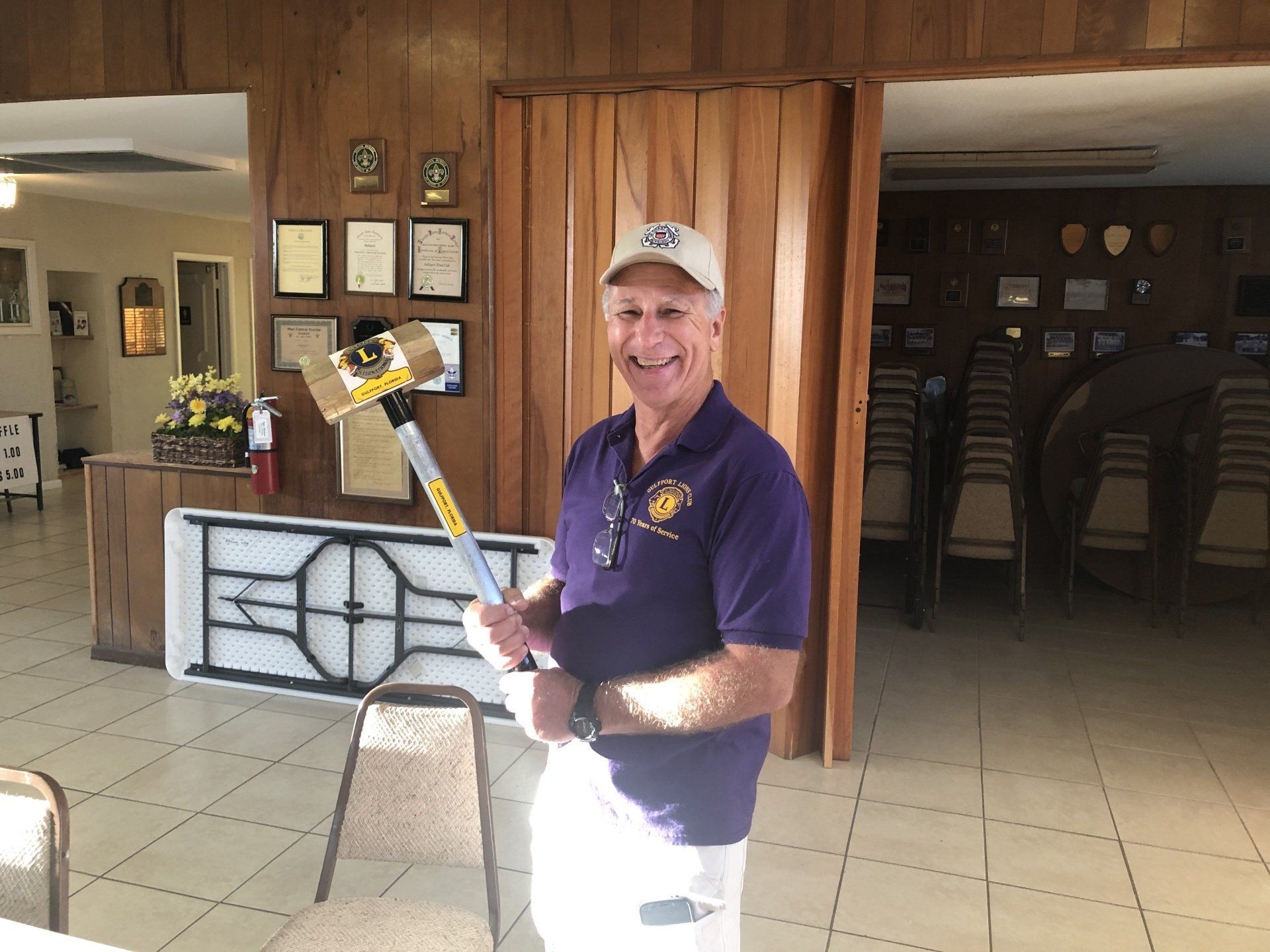 Man in purple shirt and cap holds a large mallet indoors, smiling.