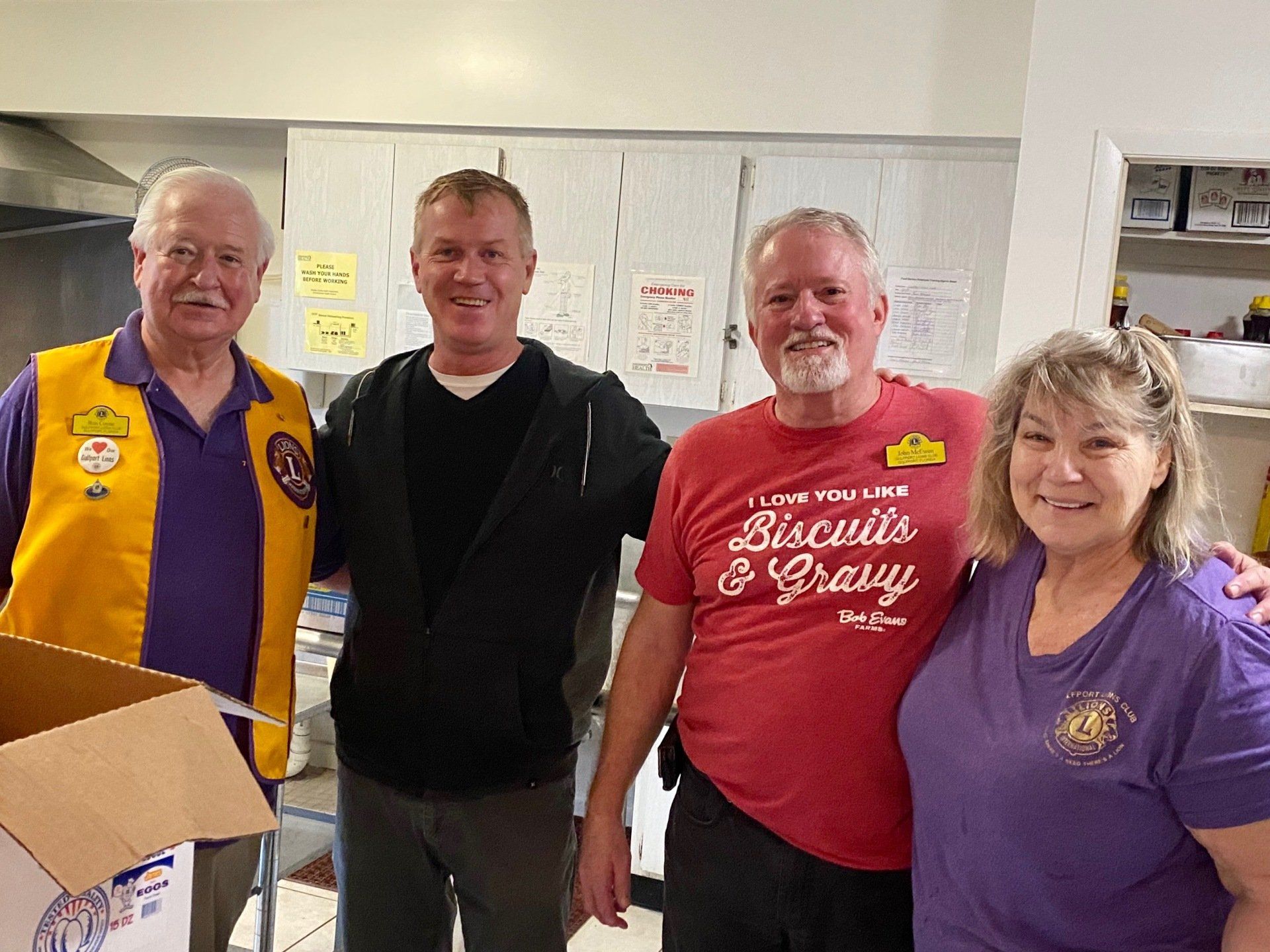 Four people in a kitchen, smiling. Two men wear Lions Club vests. One man has a 