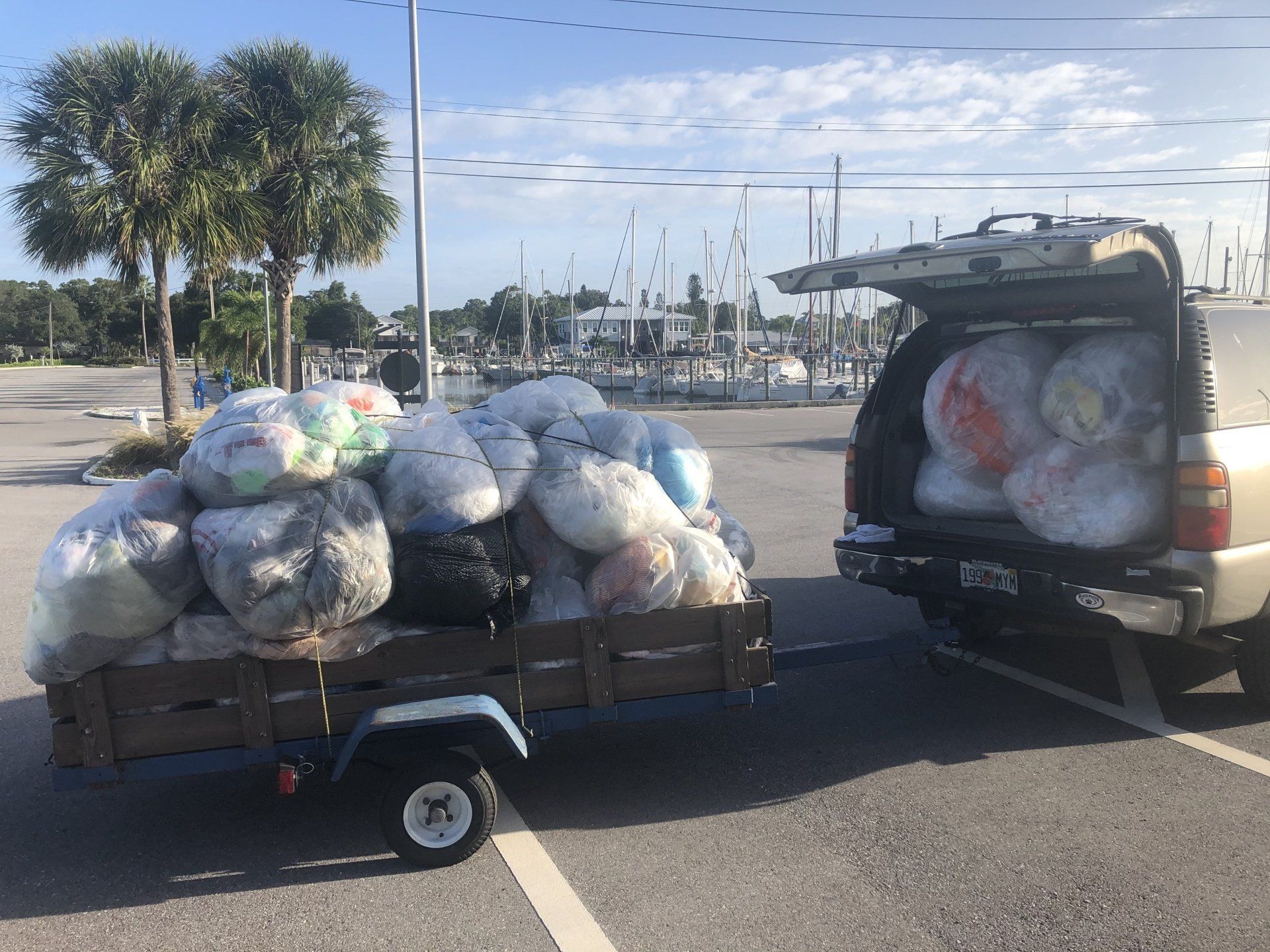 A truck towing a trailer filled with large bags, likely trash, in a parking lot near a marina.