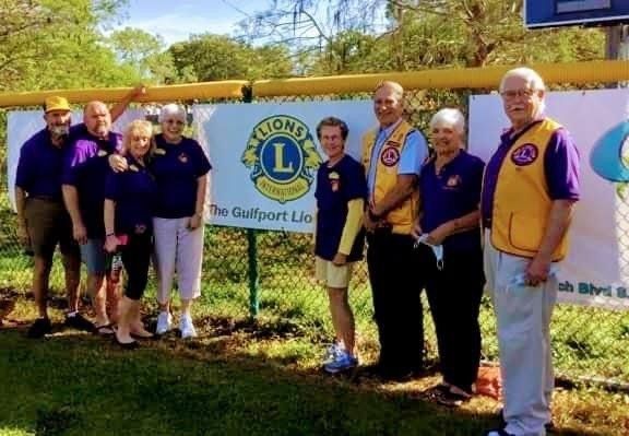 Group of Lions Club members in purple and yellow shirts stand in front of a banner in Gulfport.