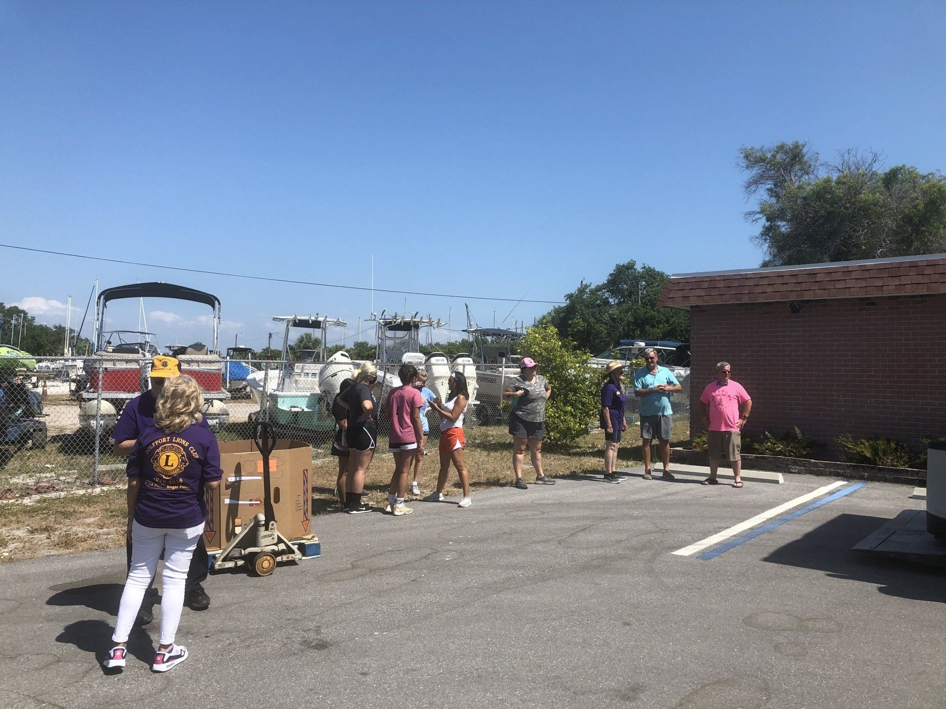 Group of people standing in a parking lot near boats on a sunny day.