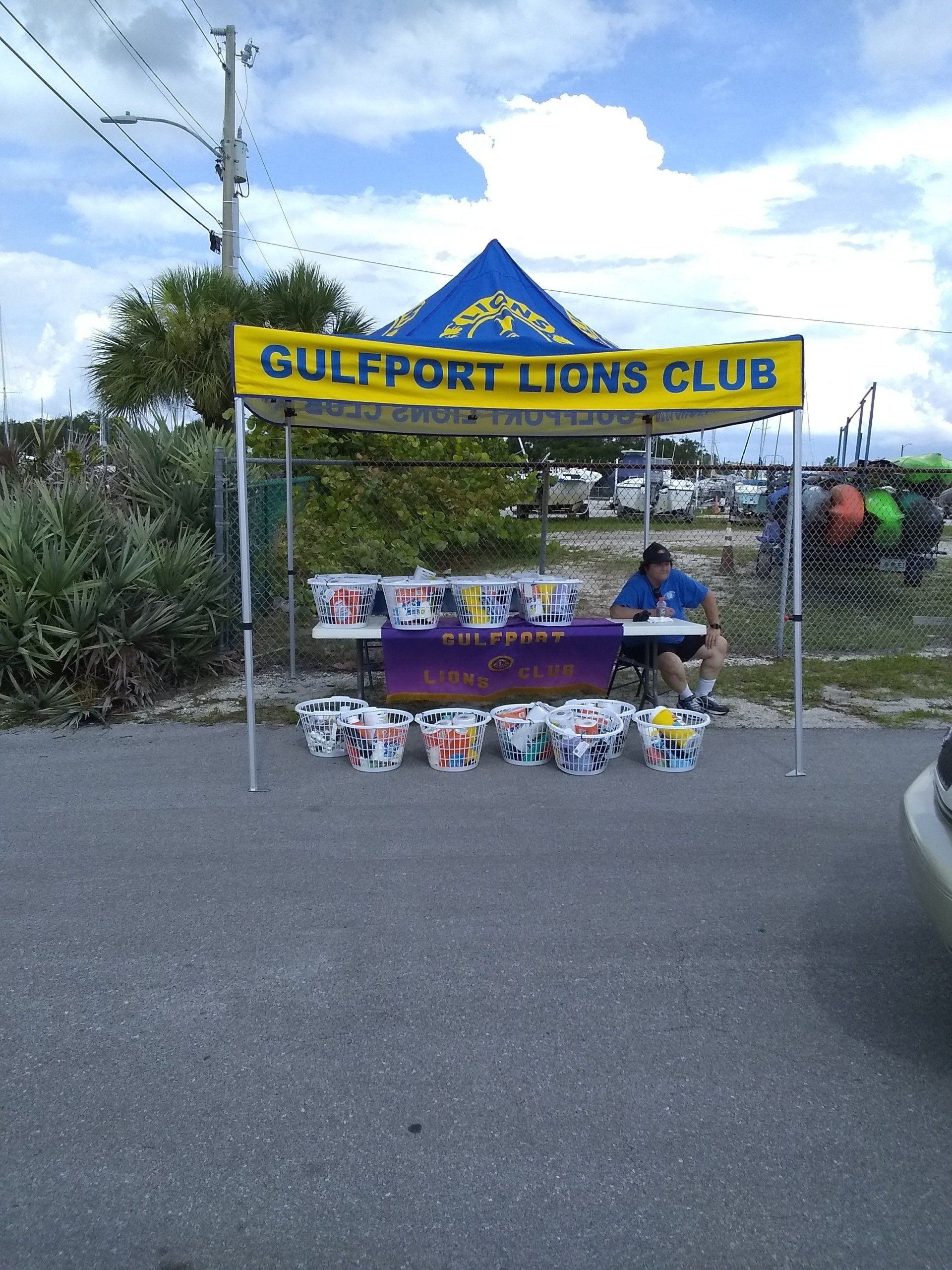 Gulfport Lions Club booth selling items; person sitting at table under yellow and blue canopy.