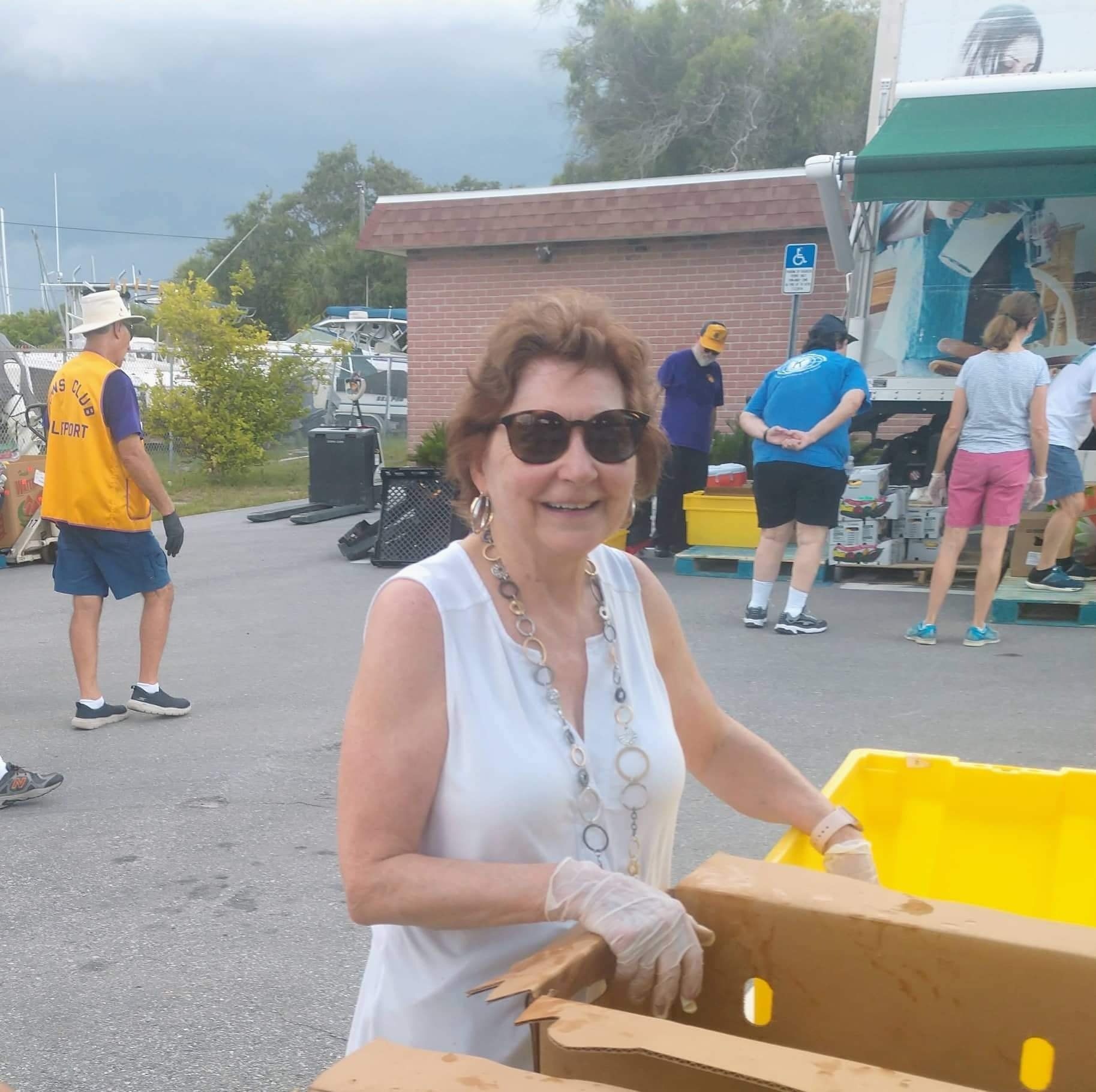 Woman in sunglasses smiles while sorting items at an outdoor event. Others assist nearby.