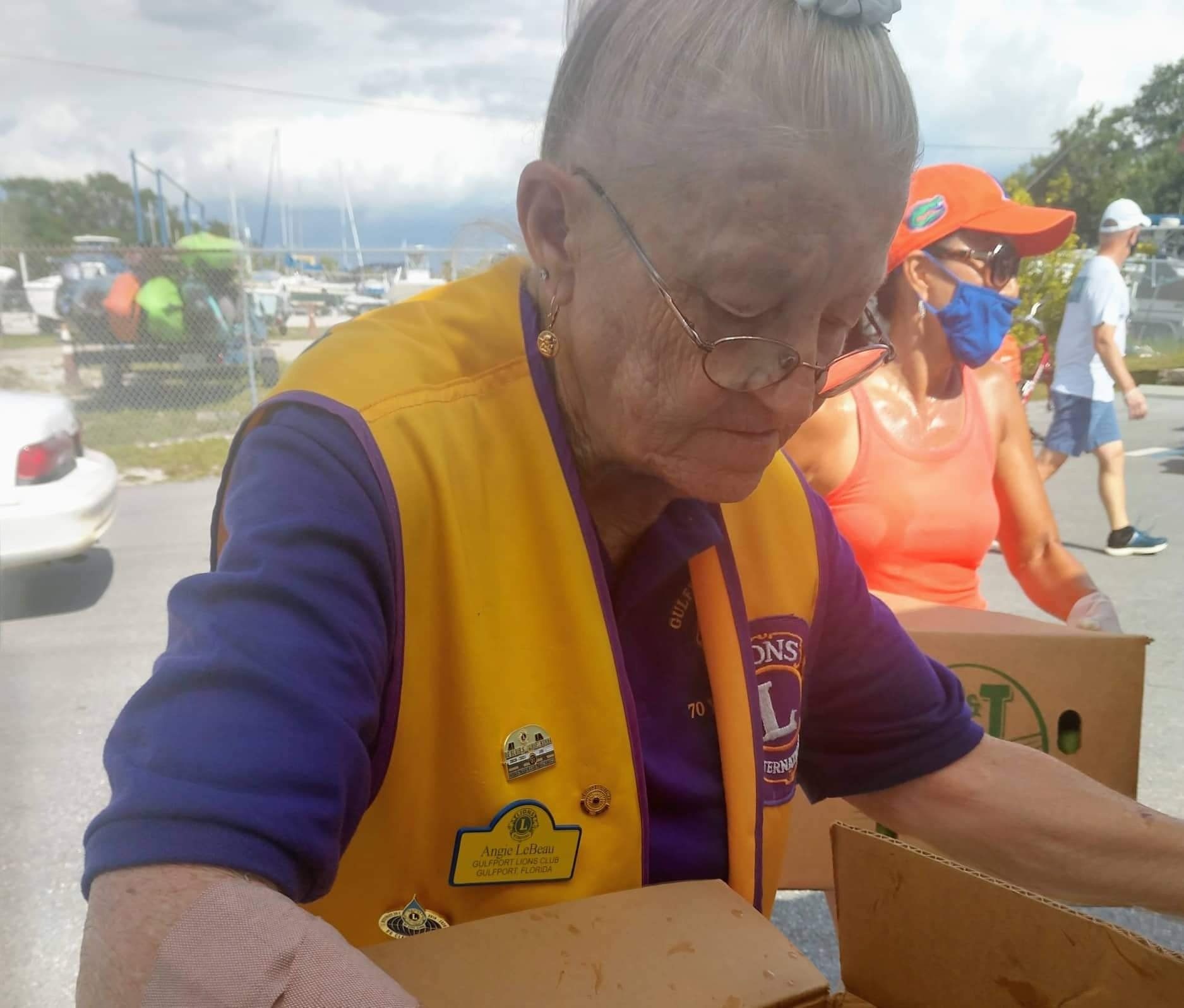 Elderly woman in Lions Club vest sorting food into boxes outdoors. Others in background.