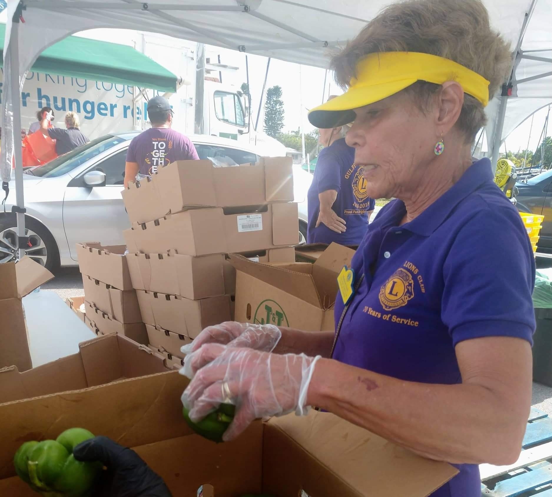 Volunteer wearing purple shirt and visor, packing produce into boxes at an outdoor food distribution site.