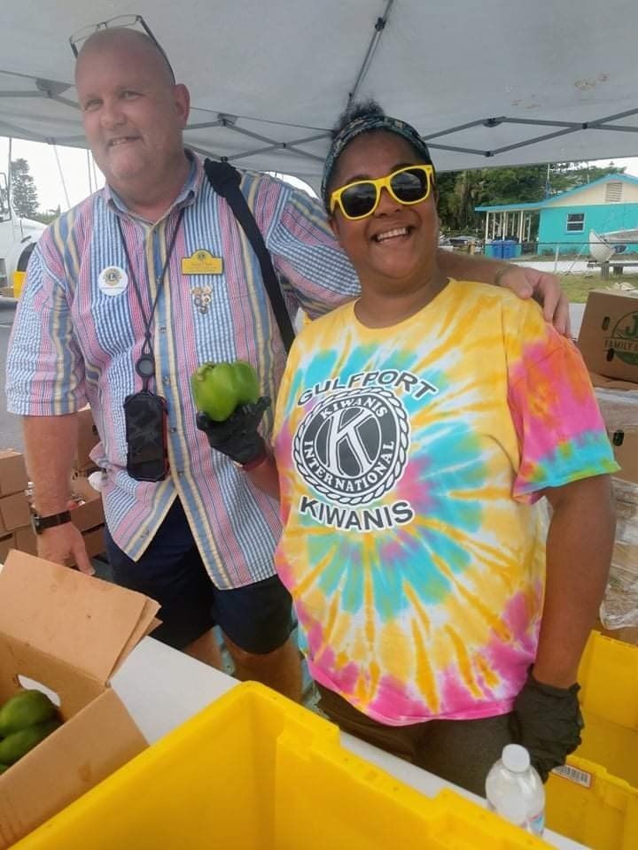 Two people smiling at a food stand; man with striped shirt and woman with tie-dye, holding a green pepper.