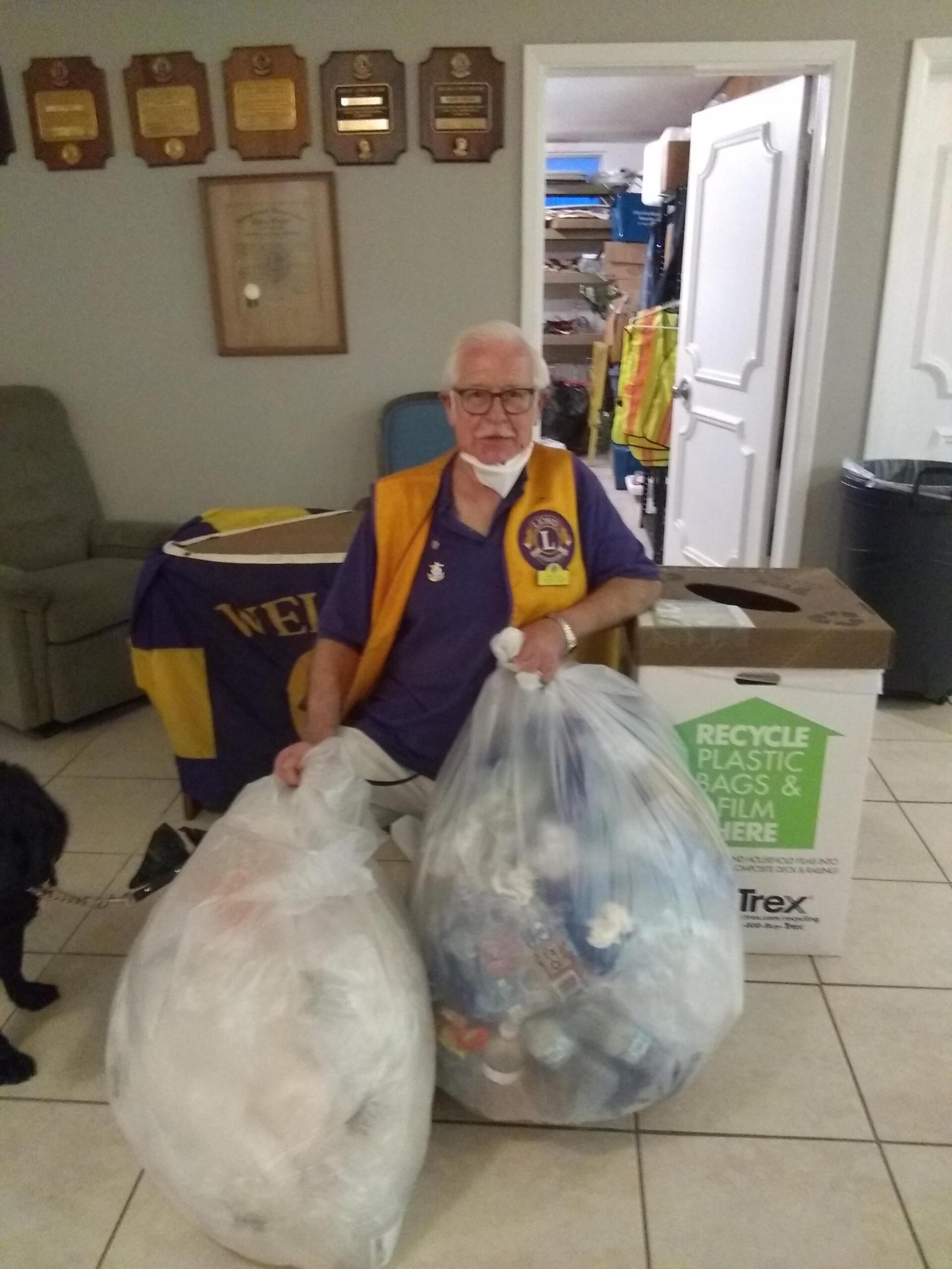 Man in Lions Club vest holding two bags of plastic recyclables near a recycling box.
