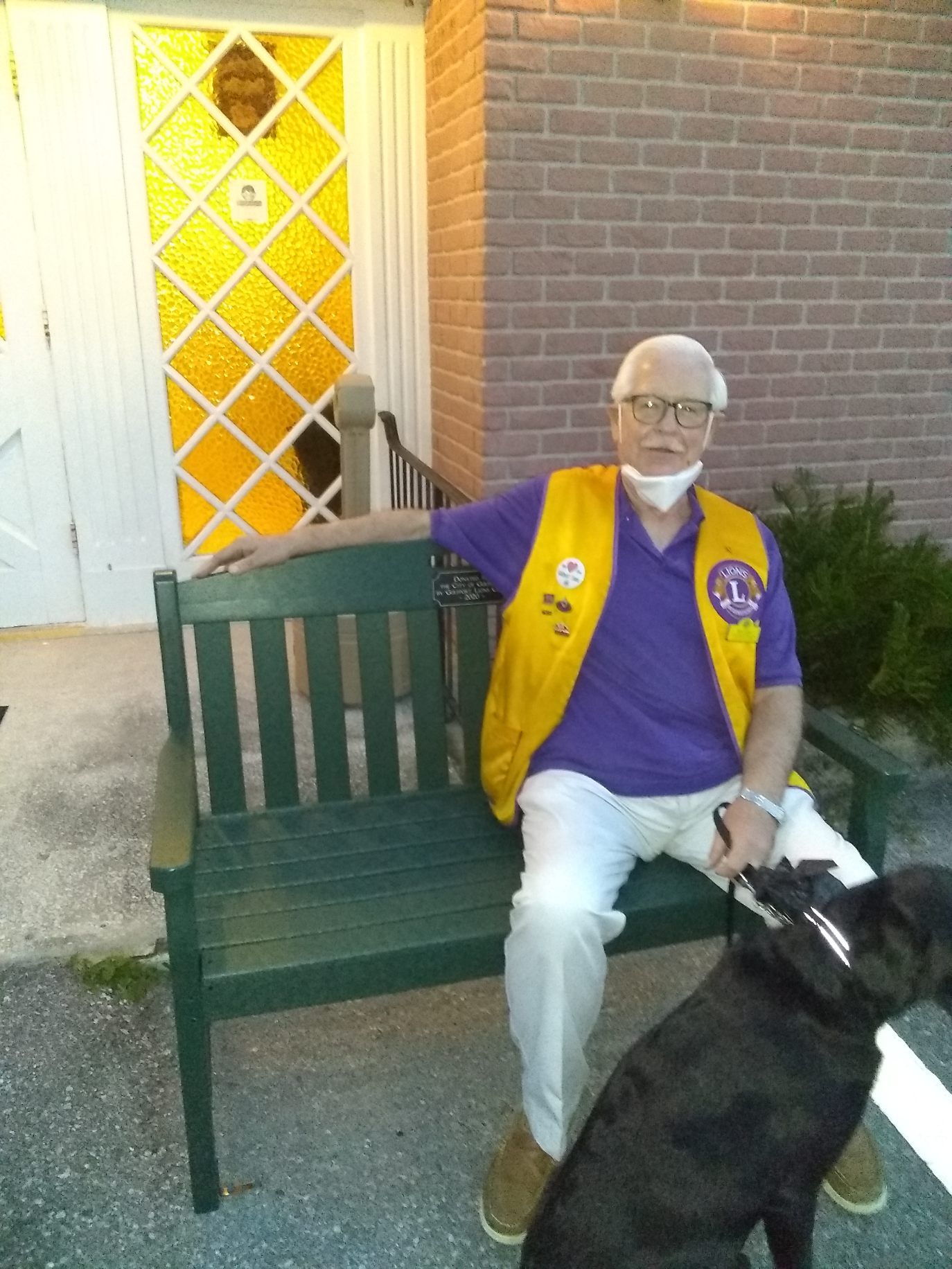 Man in Lions Club vest sits on a green bench with a black dog, near a building with yellow stained-glass window.