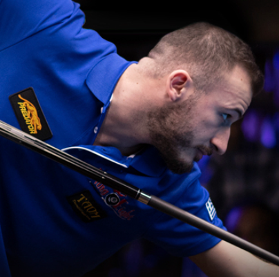 A person's hand holding a cue stick about to strike a white billiard ball on a green pool table, with a blue ball nearby.