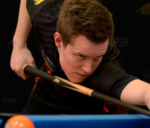 A dimly lit pool hall with several green felt tables, overhead lamps, and a rack of cues.