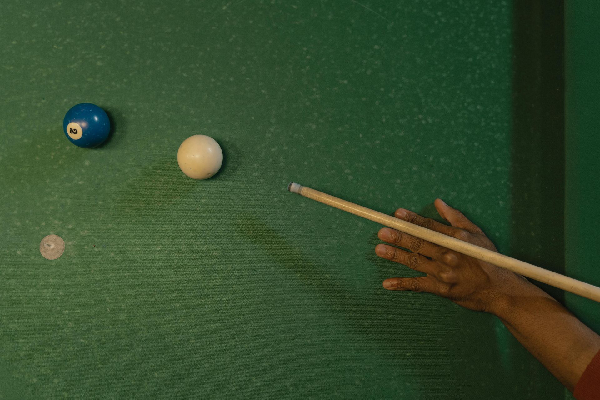 A person's hand holding a cue stick about to strike a white billiard ball on a green pool table, with a blue ball nearby.