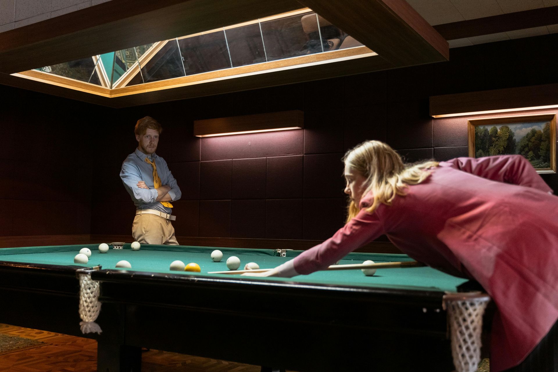A woman in a red coat plays pool while a man watches with a pool cue in a dimly lit room.