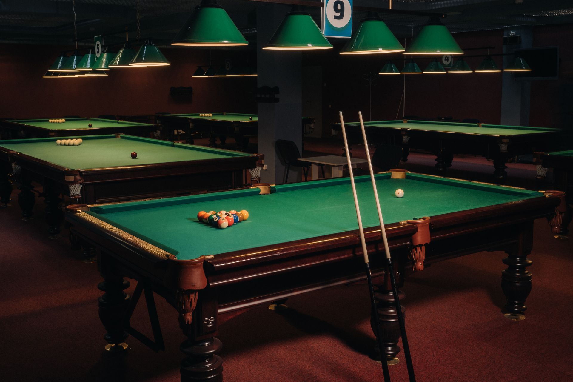 A dimly lit pool hall with several green felt tables, overhead lamps, and a rack of cues.