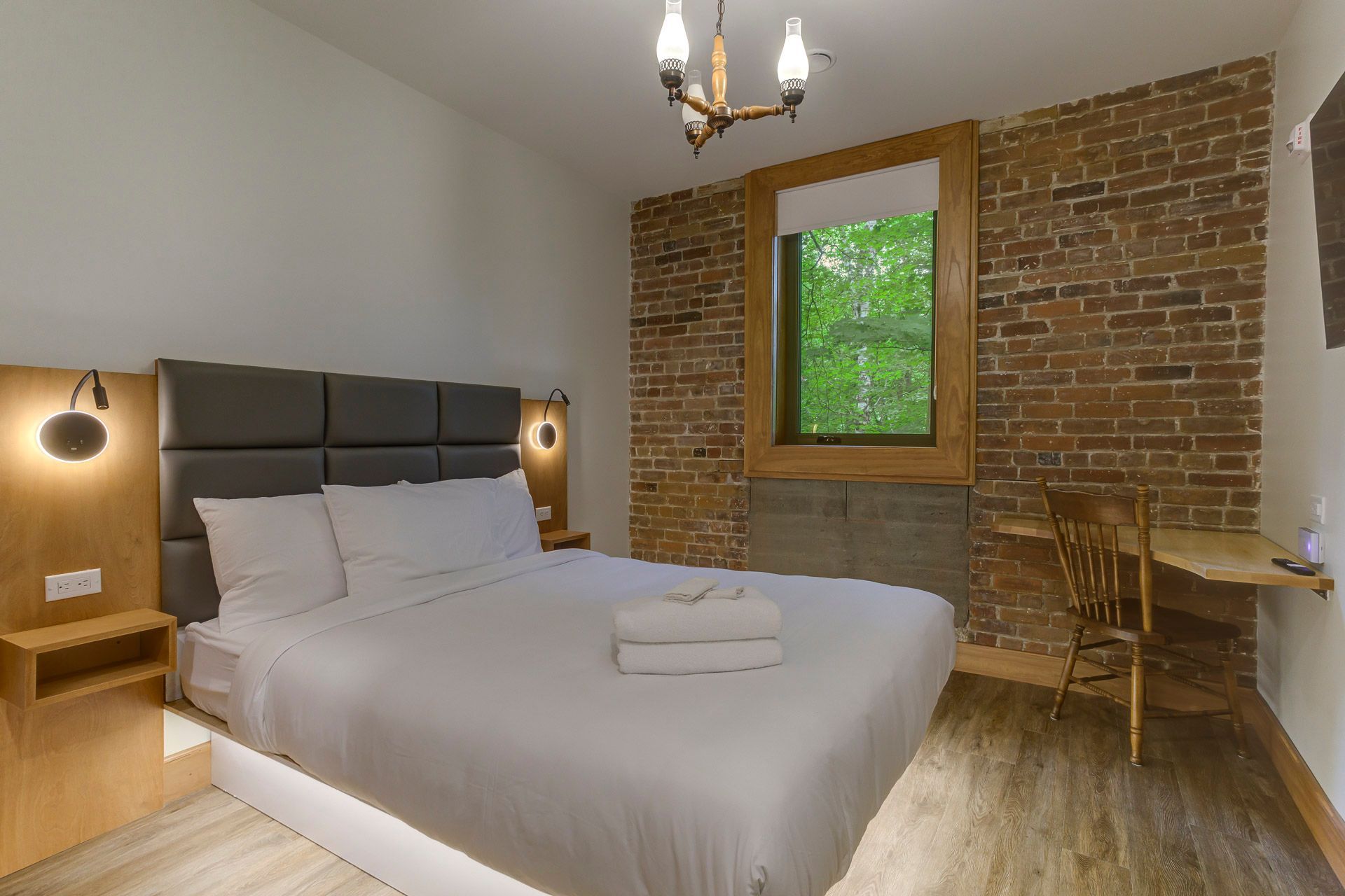 Chambre d'hôtel avec un lit, un mur en briques apparentes, un bureau en bois et une fenêtre avec des arbres à l'extérieur.
