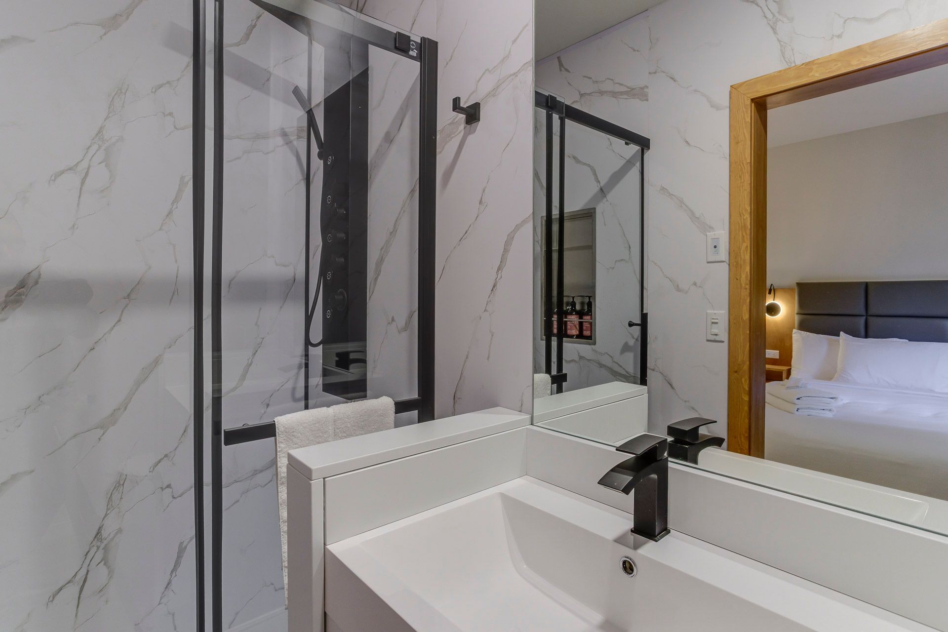 Salle de bain moderne avec murs en marbre blanc, douche à cadre noir, lavabo blanc et miroir reflétant un lit.