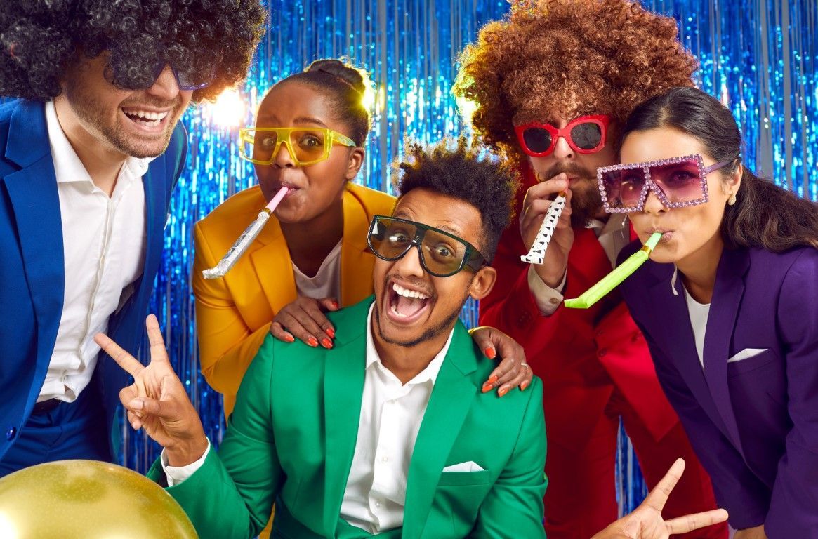 Four people in colorful suits and glasses pose joyfully at a neon party with party blowers.