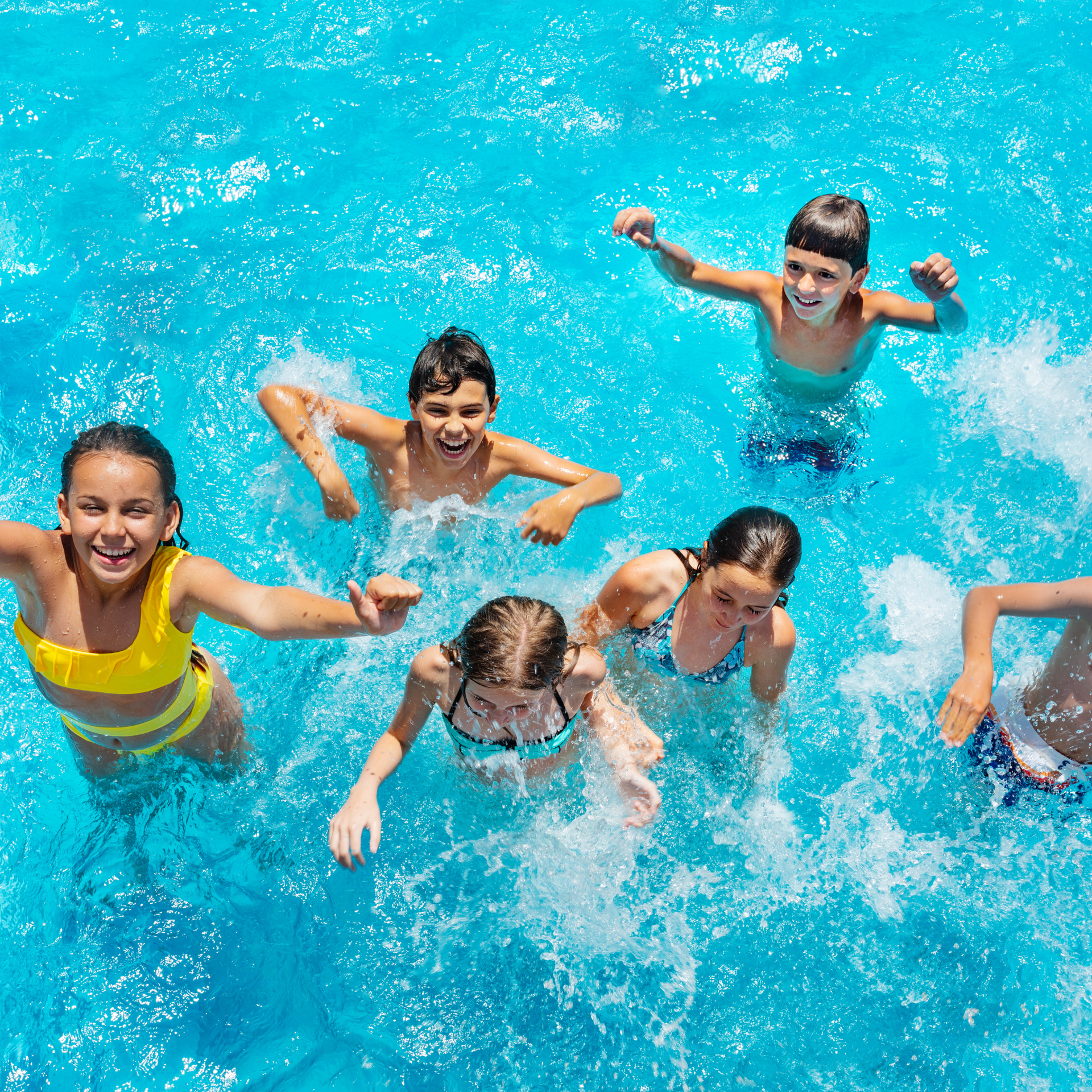 A group of children smiling and playing in bright blue swimming pool water, surrounded by splashes.