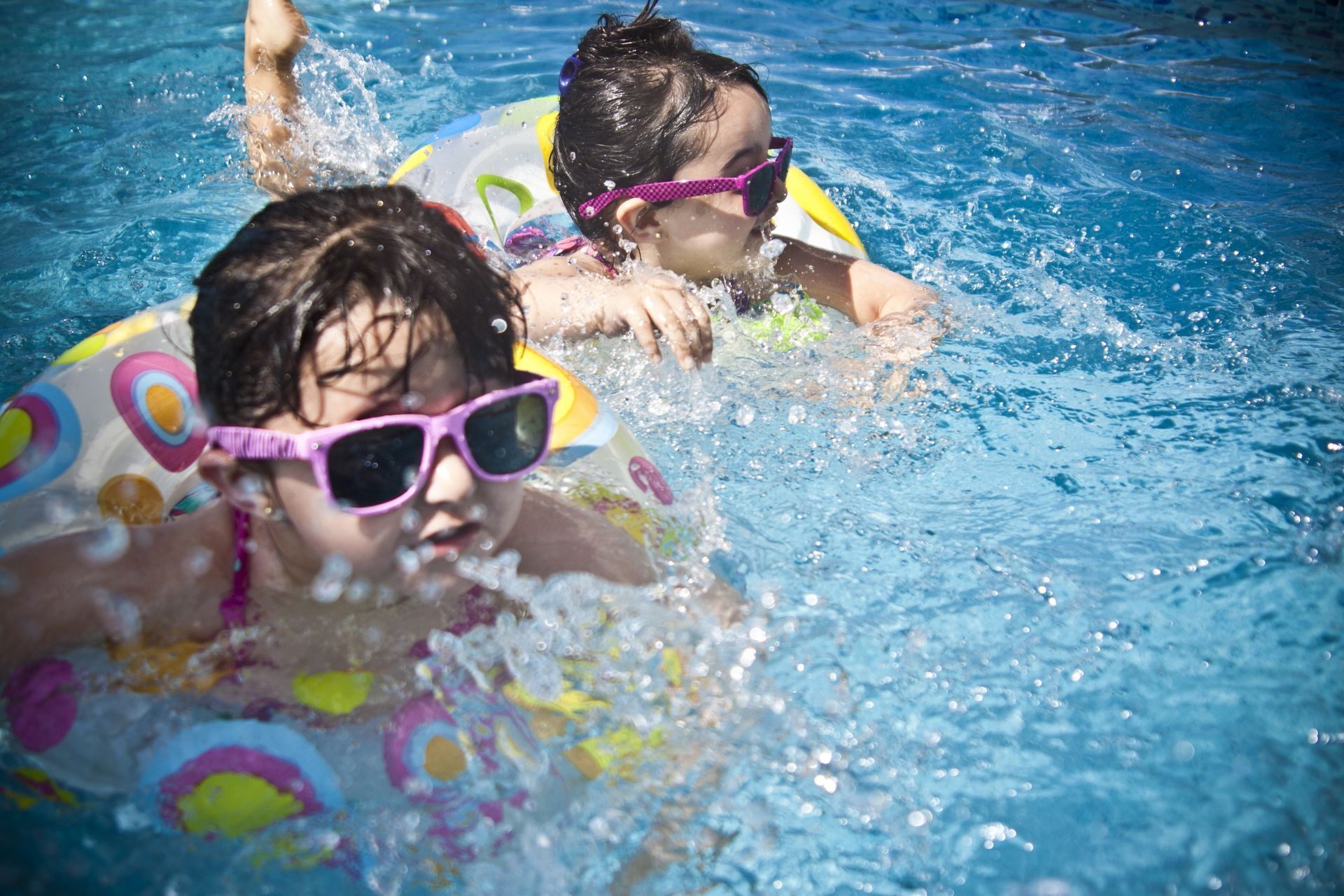 People lounge on colorful inflatable pool floats in a backyard swimming pool, with fruit-shaped towels on the lawn.