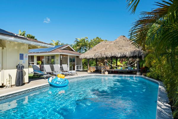 A swimming pool with a blue float, patio chairs, and a thatch-roofed gazebo under a clear blue sky.
