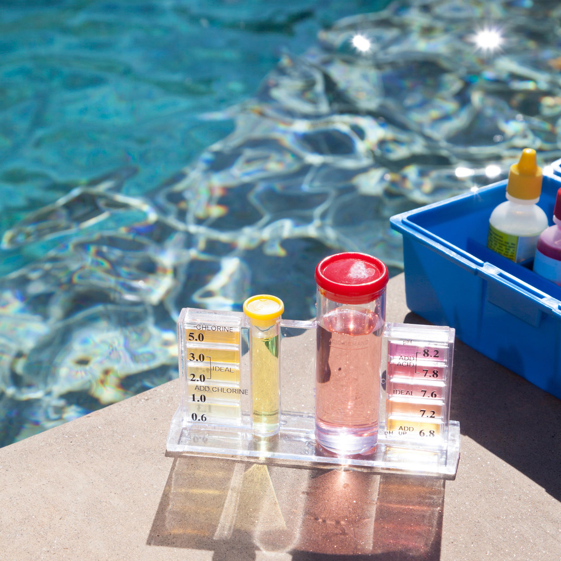 A pool water test kit sits on a deck beside a blue pool, showing yellow and pink testing liquid in clear vials.