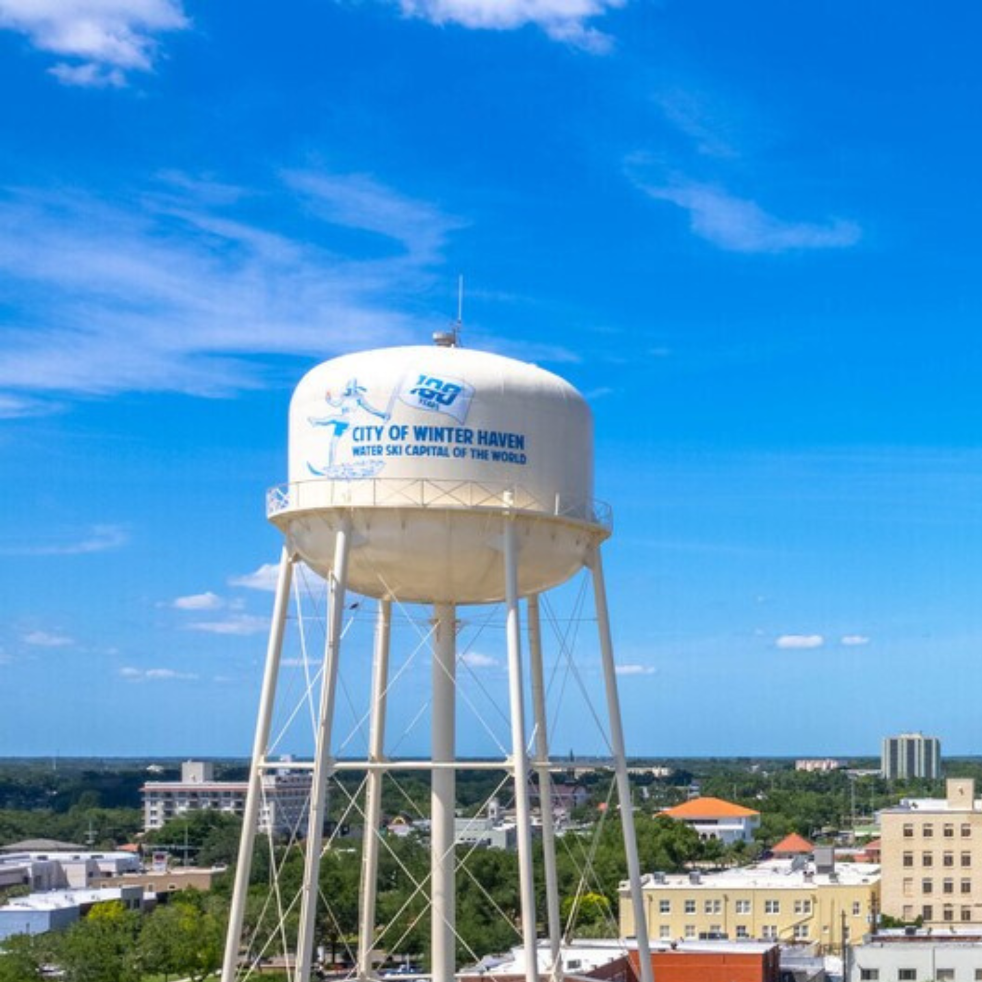 A large white water tower stands against a bright blue sky, overlooking a town with various buildings and green trees.